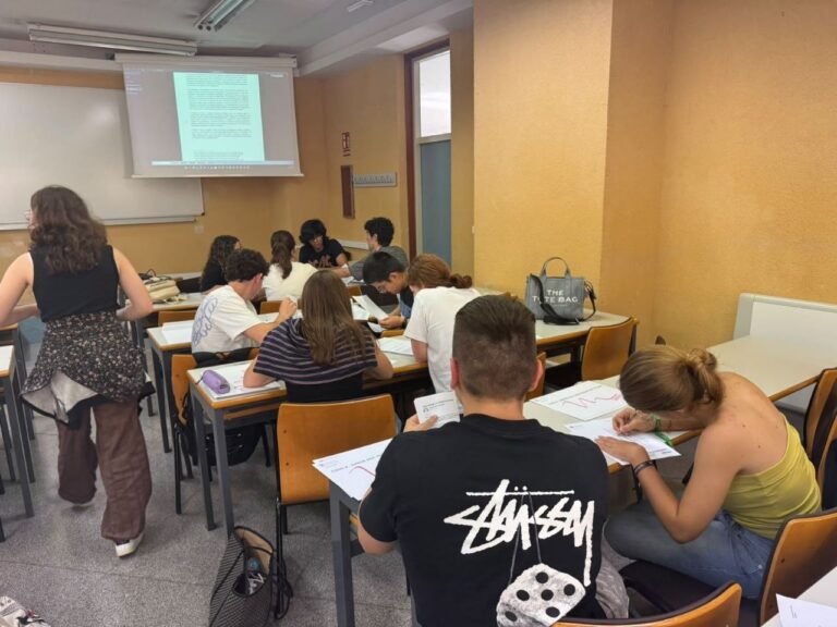 Estudiantes sentados en pupitres trabajan con documentos mientras se proyecta una presentación al fondo del aula.