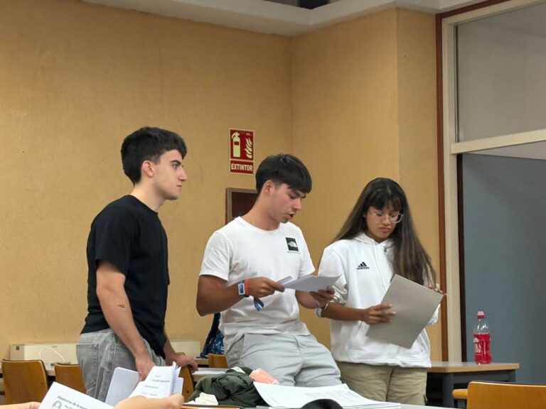 Tres estudiantes de pie revisan documentos impresos durante una actividad en el aula.